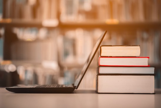 Auf einem Tisch befinden sich ein Laptop. Dahinter liegt ein Stapel mit 4 Büchern. Im Hintergrund ein großes Bücherregal, mit vielen Büchern.