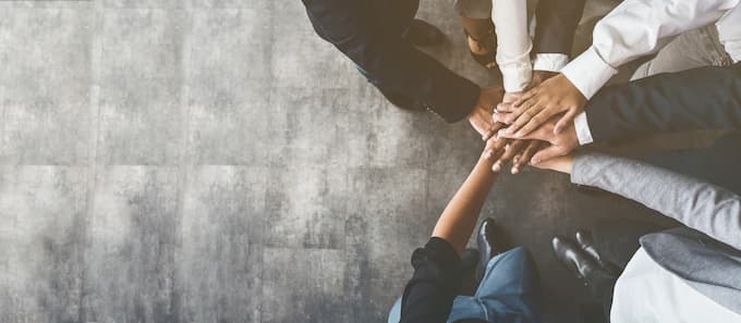 7 unterschiedliche Menschenhände stehen im Kreis und legen ihre Hände zusammen.